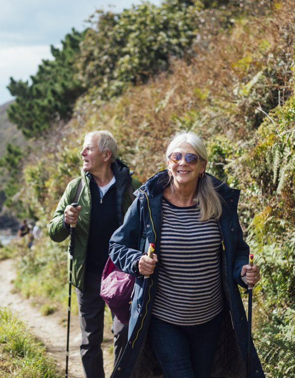 A senior couple using hiking poles, hiking uphill on a coastal path in Cornwall. The woman is looking at the camera while the man is looking at the view.